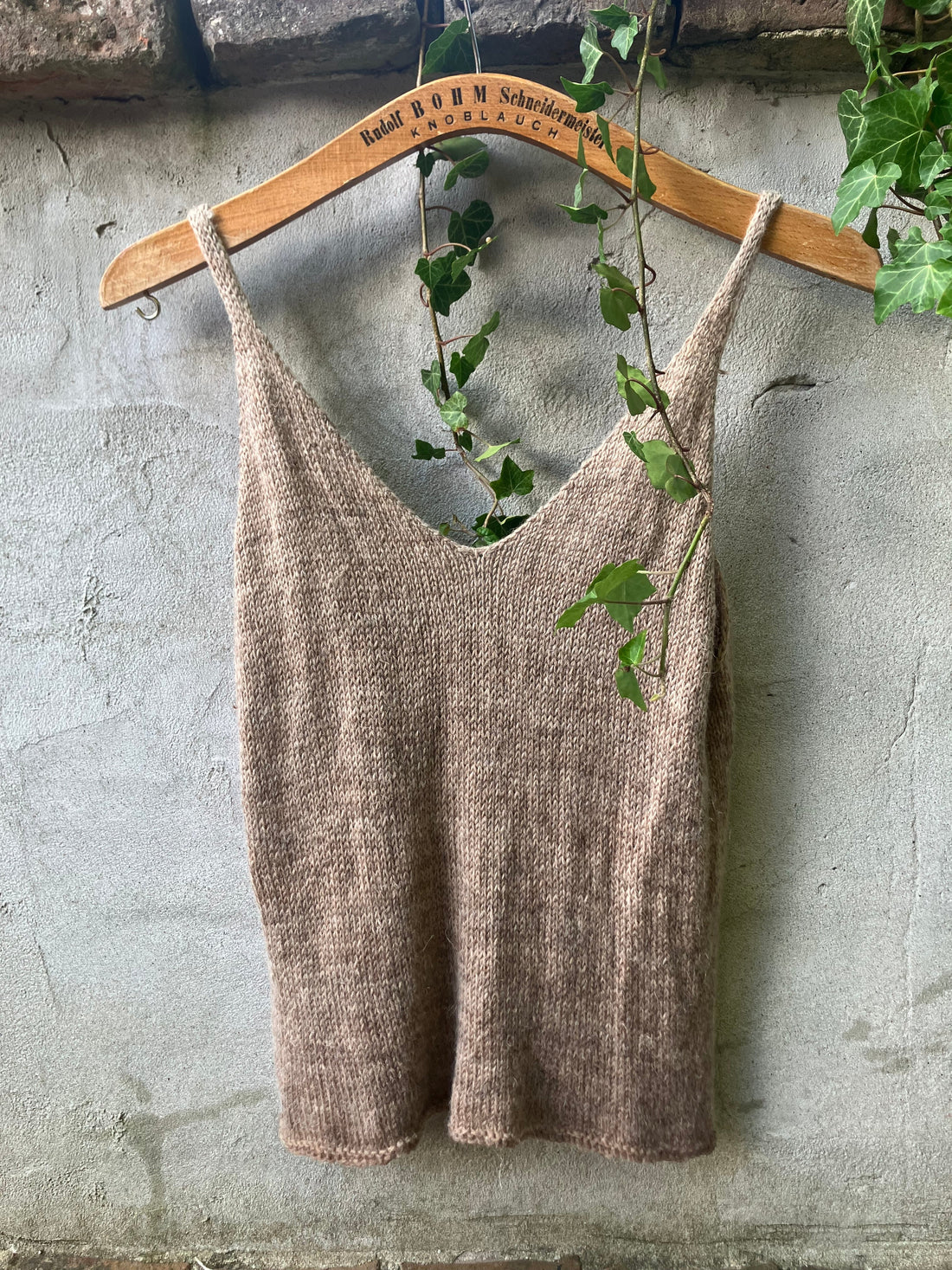A natural beige coloured knitted top hanging on an old fashioned hanger in front of a roustic cement wall, ivy hanging over it. 